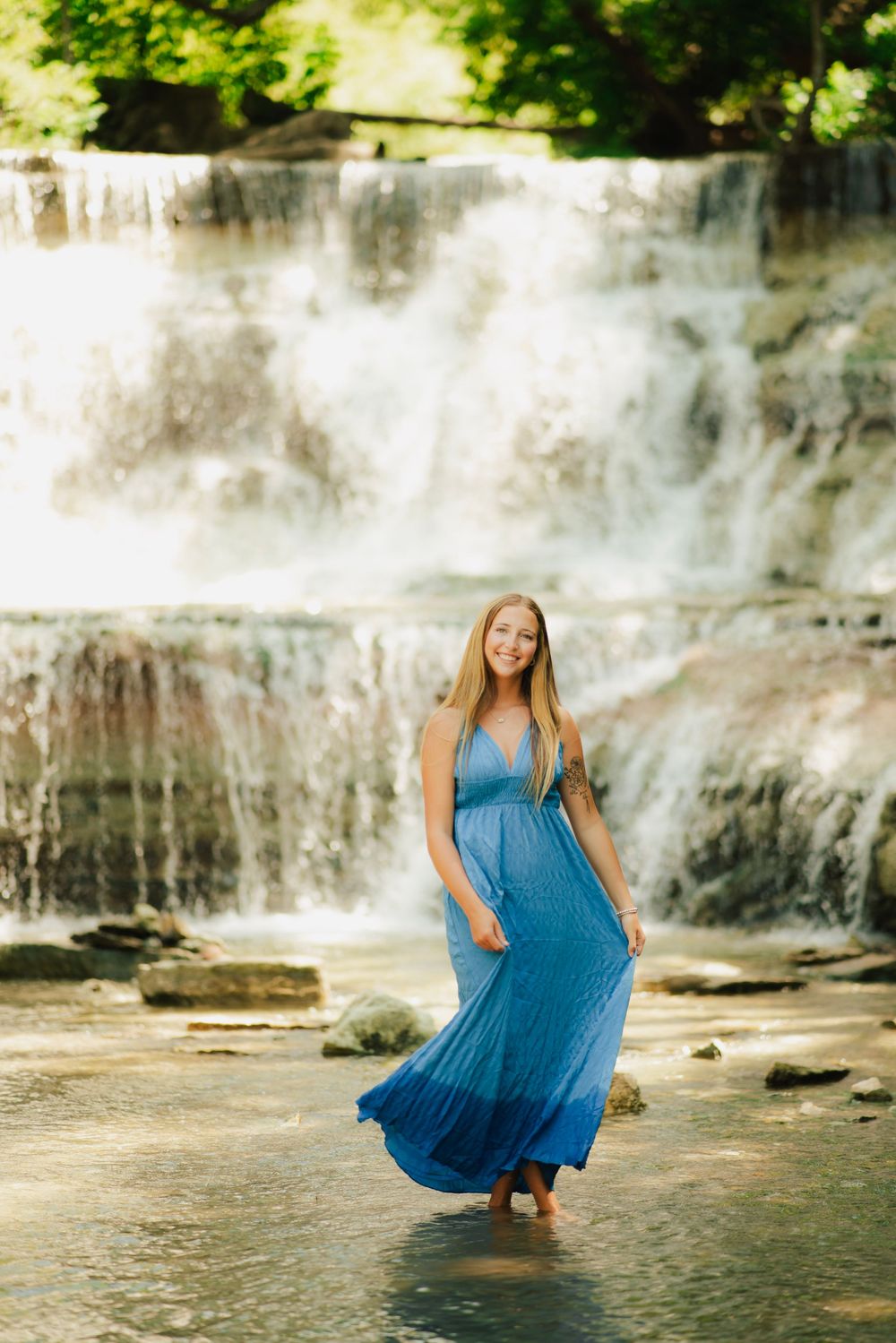 Young woman in blue maxi dress standing in shallow water before a cascading waterfall.