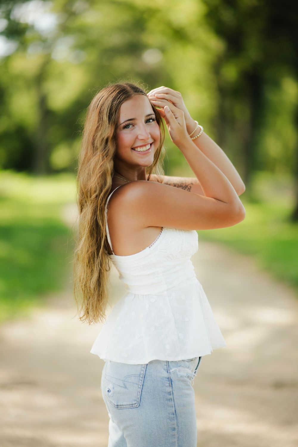 Smiling young woman in white top and jeans poses on a sunny park path with long wavy hair and a tattoo.