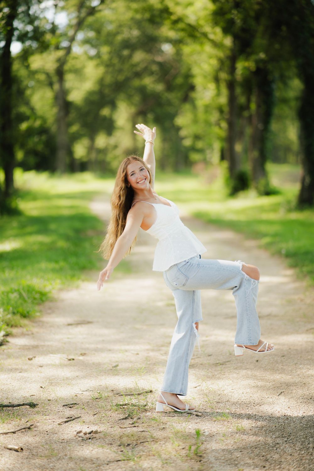 Joyful young woman in white top and ripped jeans dancing on a scenic tree-lined path in a park.