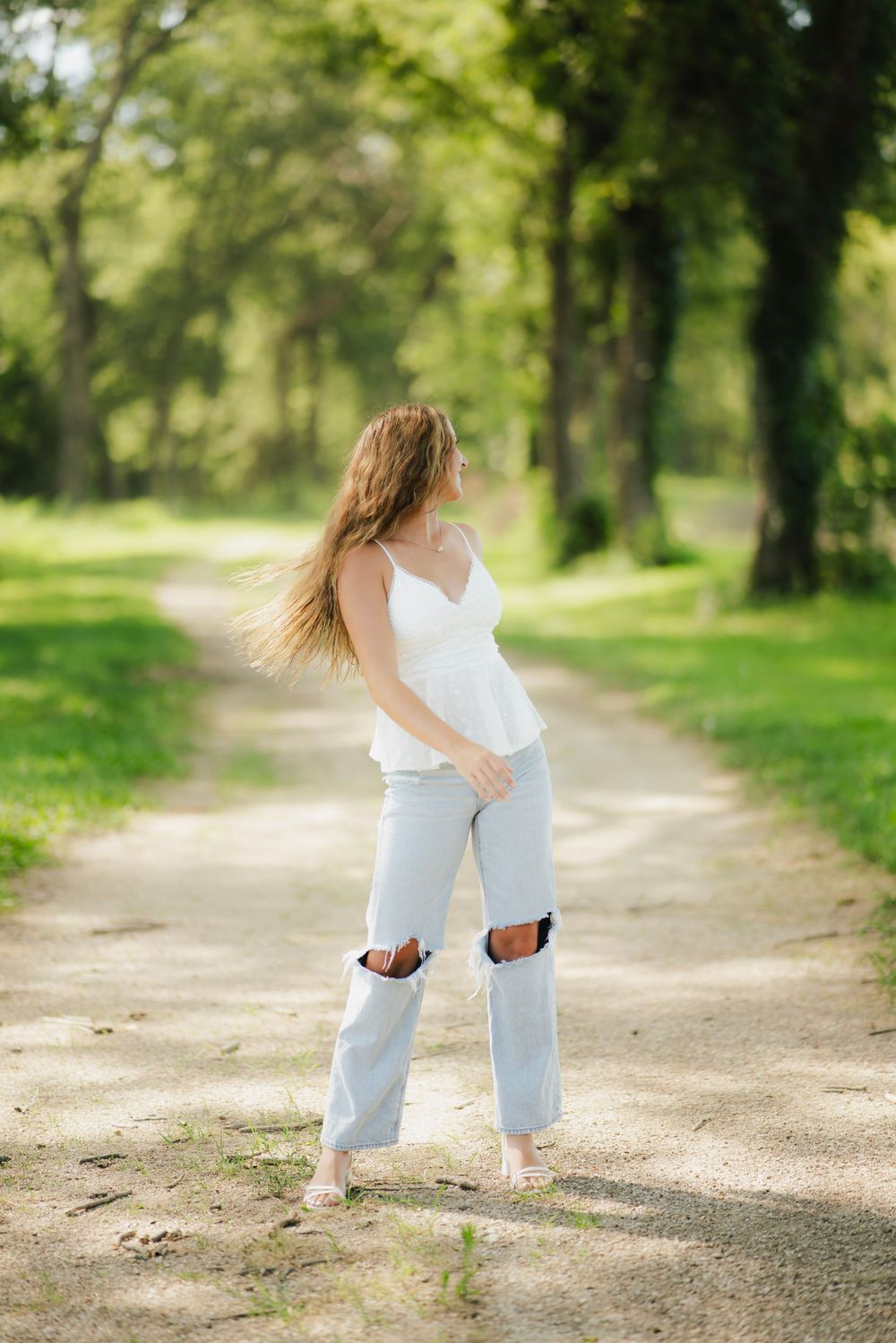Young woman in white top and ripped jeans walking on a sunny park path surrounded by lush green trees.