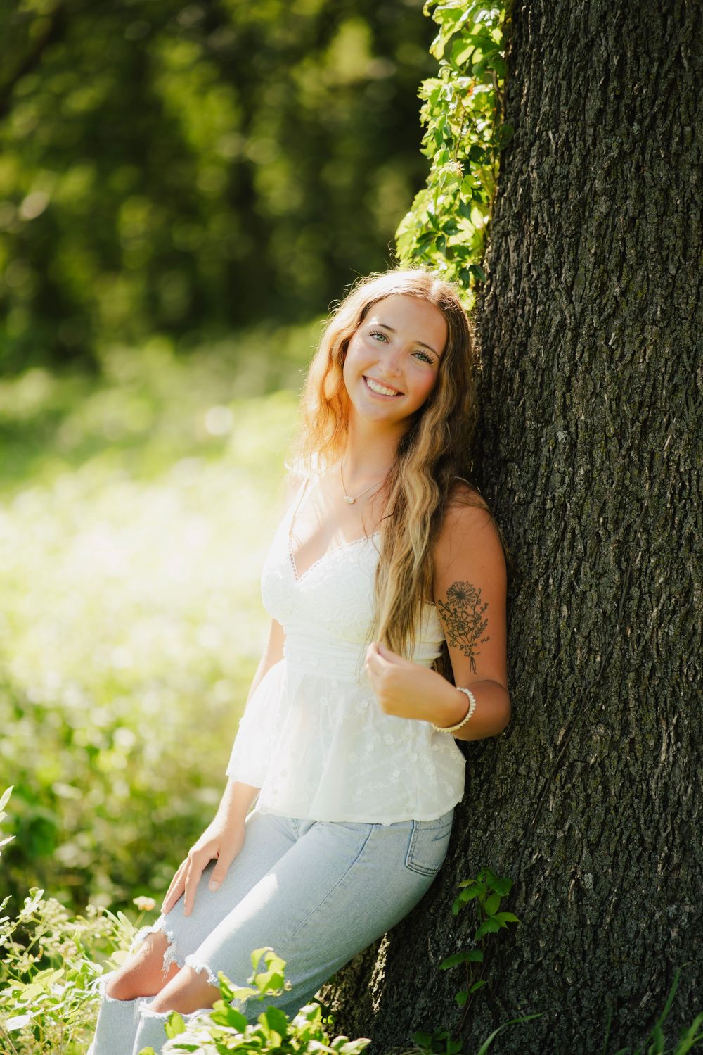 Smiling young woman with tattoo leans against large tree in sunny park wearing white lace top and jeans.