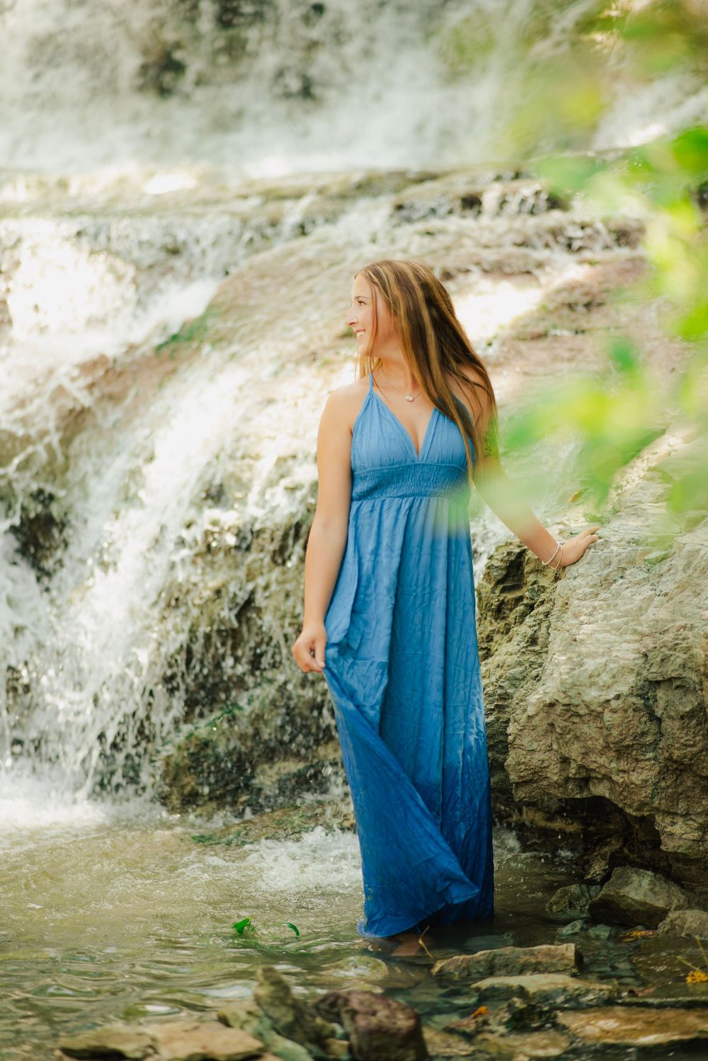 Woman in blue maxi dress standing by a waterfall on rocky terrain, with long hair and serene expression.