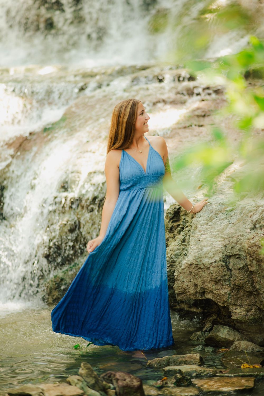 Woman in blue maxi dress standing near a waterfall on rocky terrain, smiling outdoors.