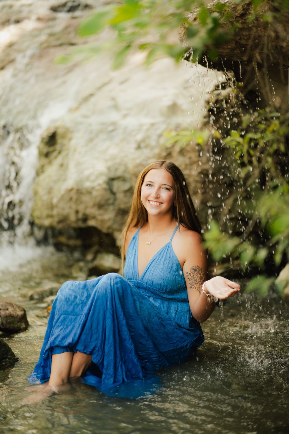 Smiling young woman in blue dress sitting in stream, playfully splashing water near a waterfall.