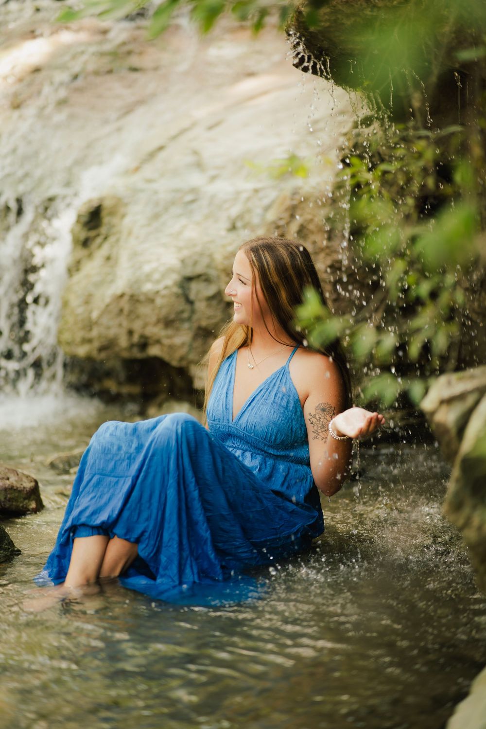 Young woman in blue dress sitting in shallow creek water beside a rocky waterfall.