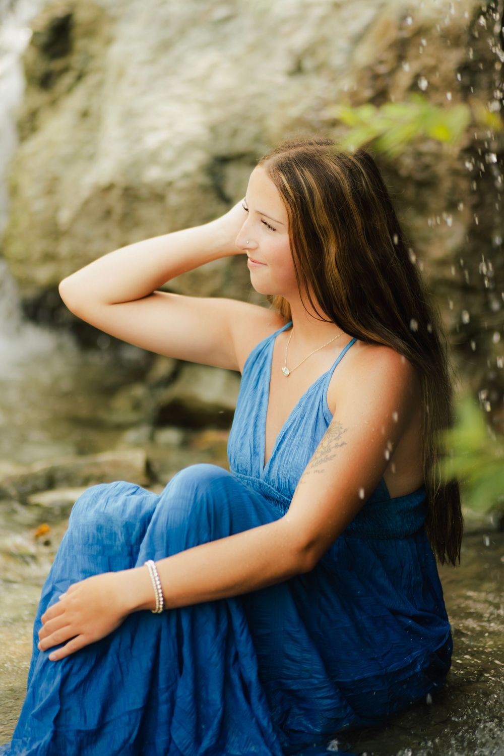 Young woman in blue dress sits near waterfall, hand in hair, tattoo visible, wearing pearl bracelet and gold necklace.