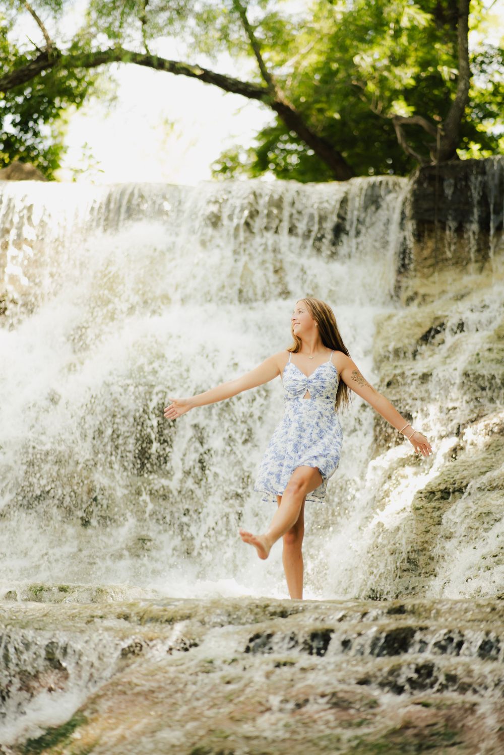 Joyful young woman in floral dress dancing freely in front of a beautiful cascading waterfall in nature.
