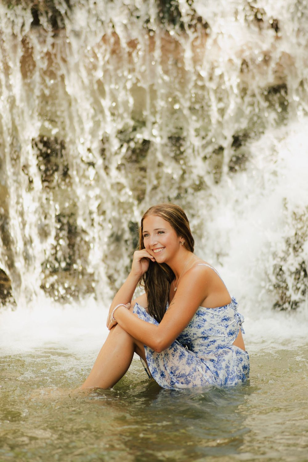 Young woman in blue floral dress sitting in shallow water before a cascading waterfall, smiling.