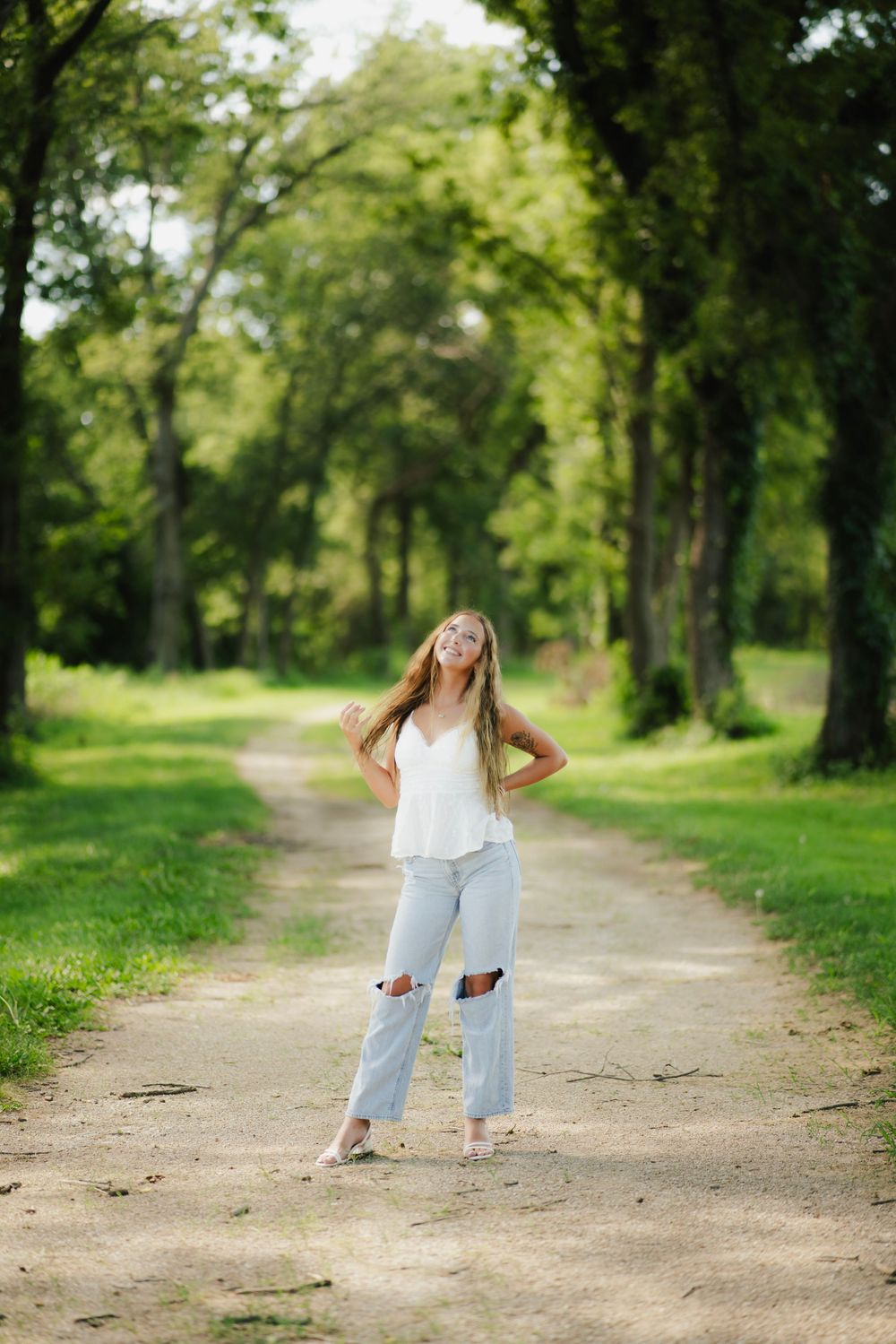 Young woman in white top and ripped jeans poses on a tree-lined dirt path in a lush green park.