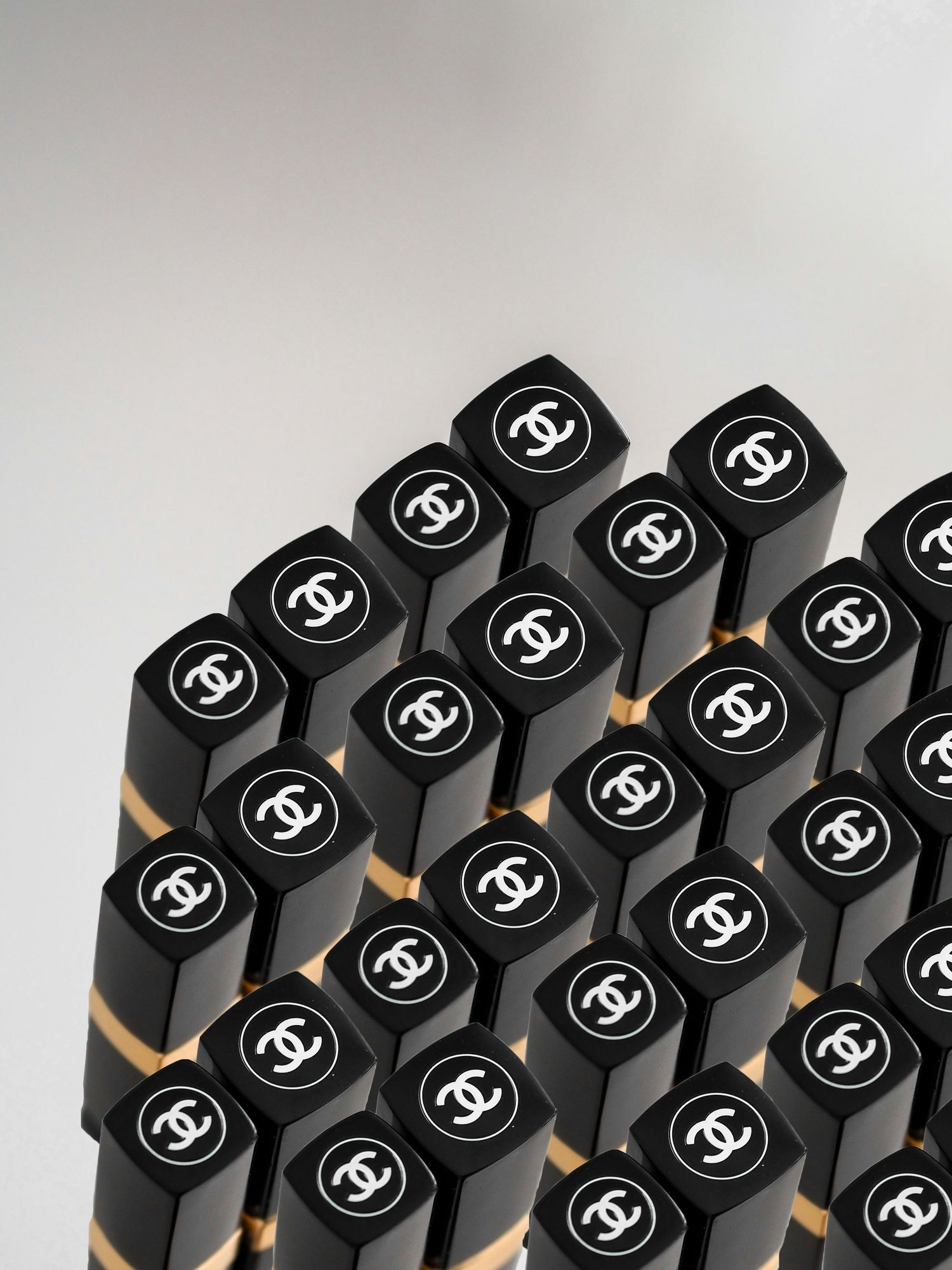 Multiple black and gold Chanel lipsticks arranged in rows, featuring the iconic CC logo on each cap.