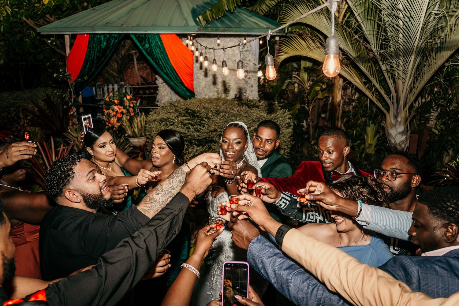 A joyful group of friends toasting with drinks at an outdoor evening celebration surrounded by lush greenery.