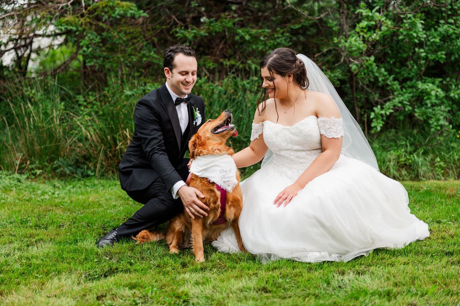 Bride and groom in wedding attire smiling while petting their golden retriever dog on a green lawn.