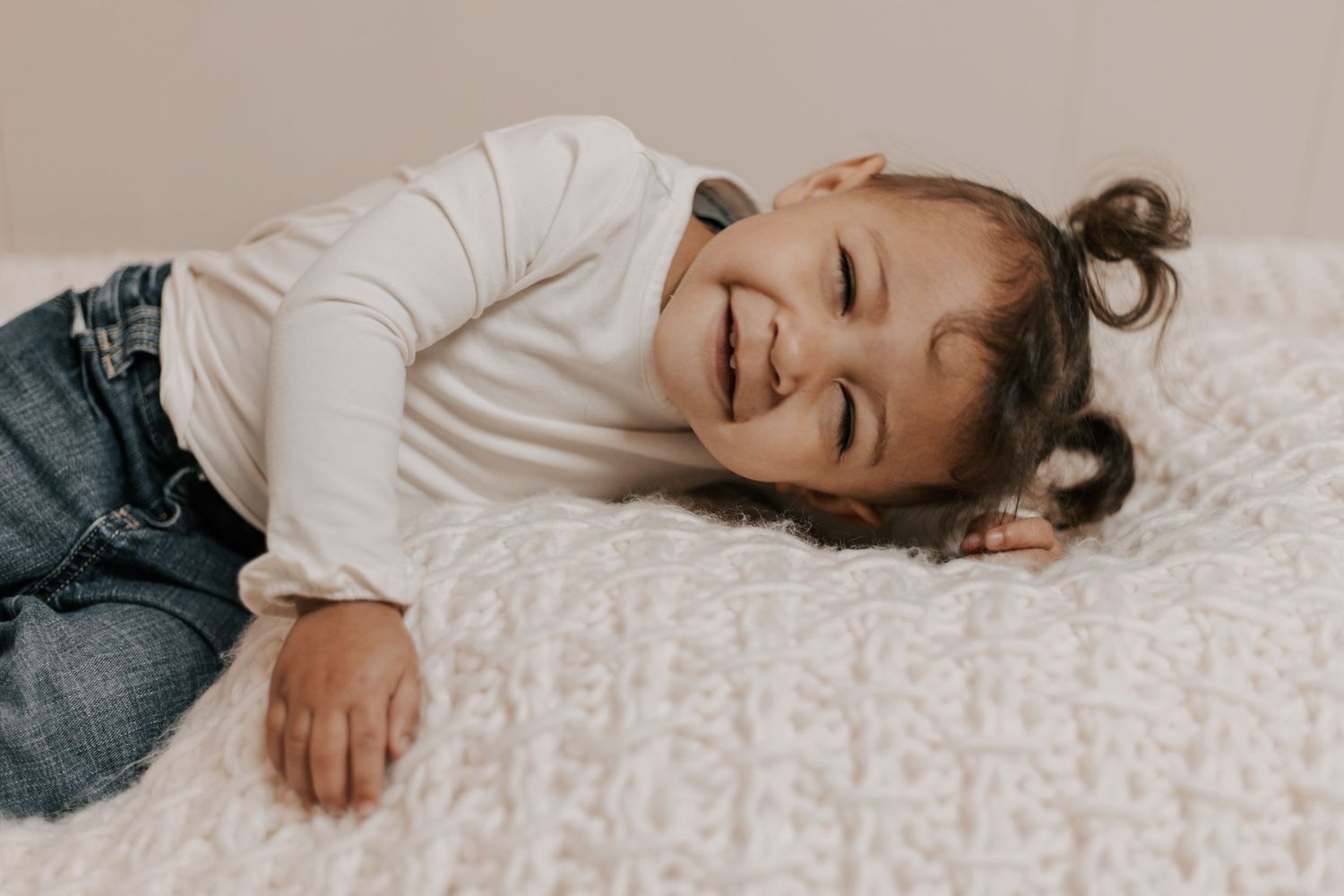 Happy little girl lying on a white knit blanket, smiling joyfully while wearing a white top and jeans.