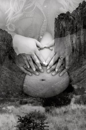 Black and white double exposure maternity photo with couple forming heart shape on pregnant belly against rocky landscape.