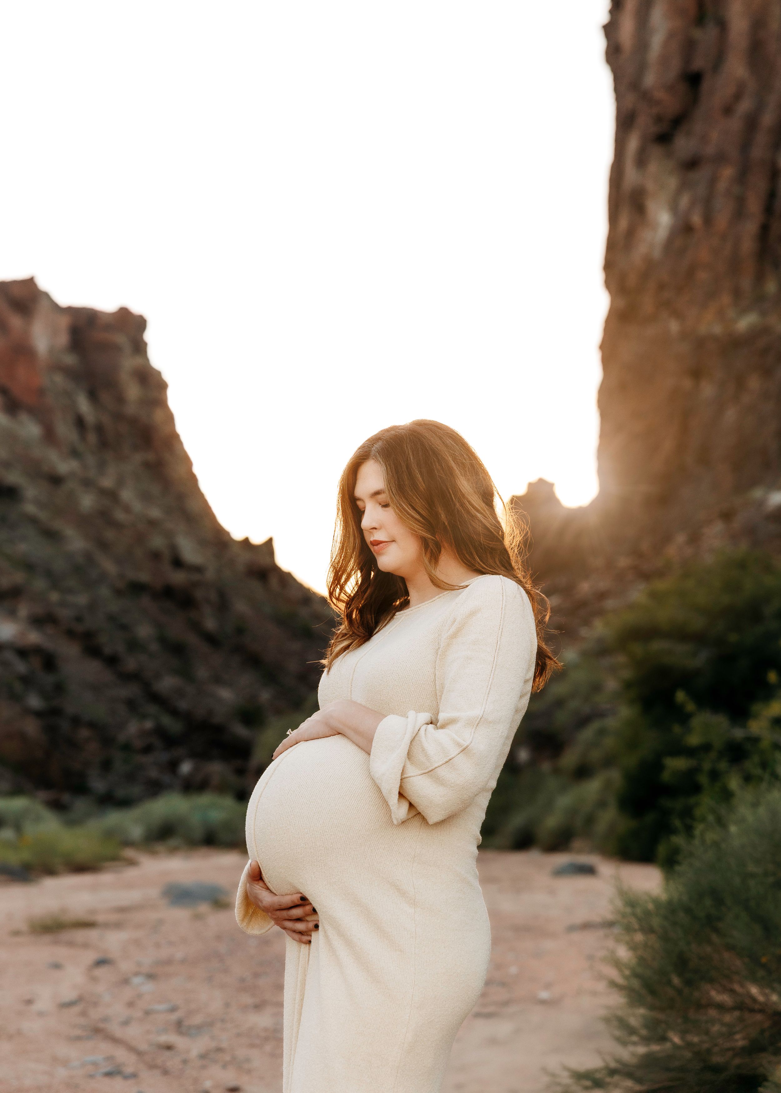Pregnant woman in white dress cradling her baby bump at golden hour among desert rock formations.