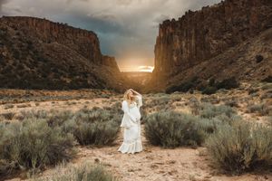 Woman in white dress standing in desert canyon landscape at sunset with dramatic storm clouds.