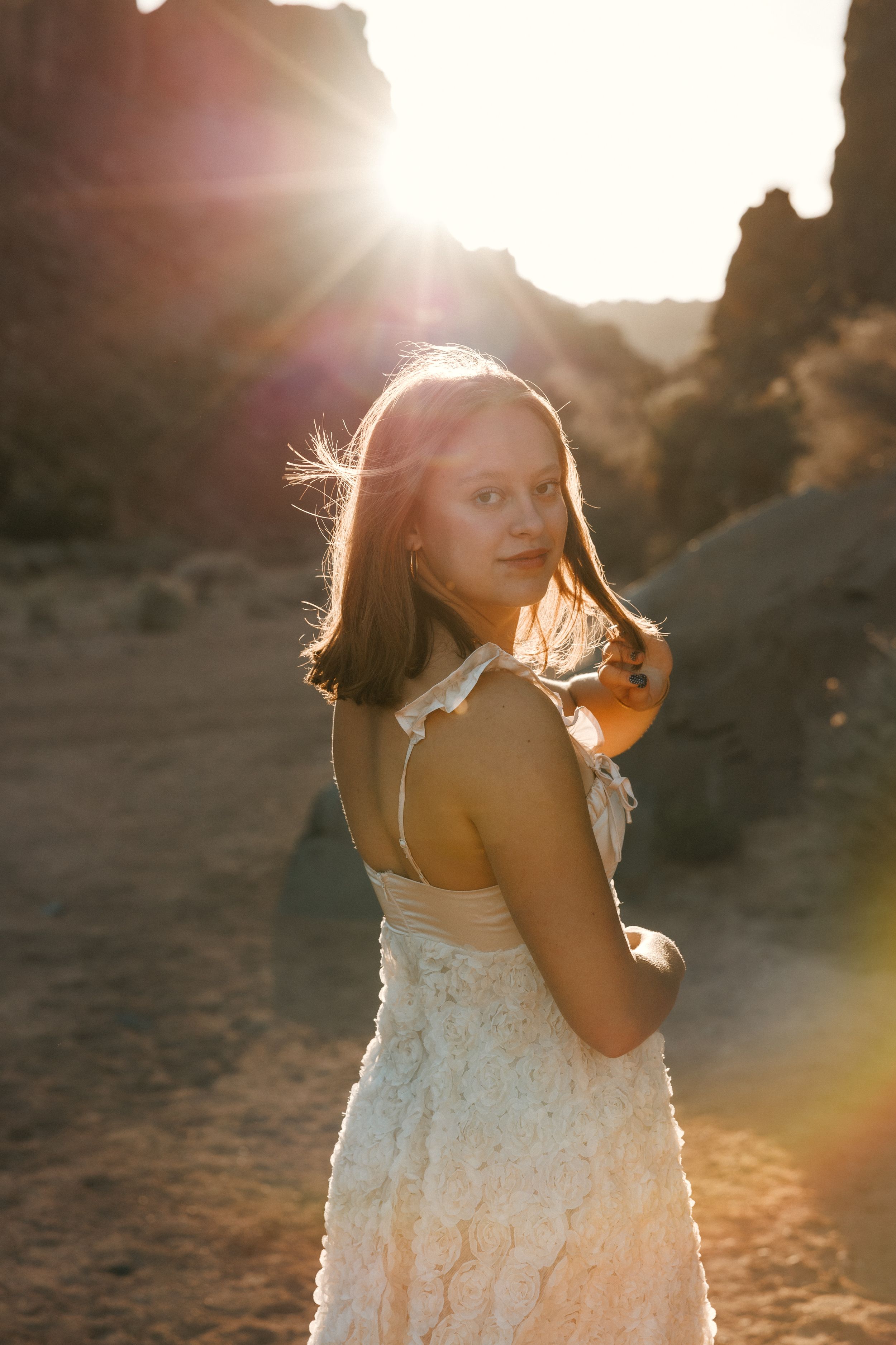 Young woman in white lace dress poses in desert canyon with golden sunset lens flare behind her.