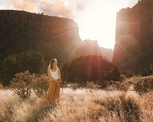 Woman in floral skirt standing in sunlit desert canyon with golden grass and rocky cliffs at sunset.