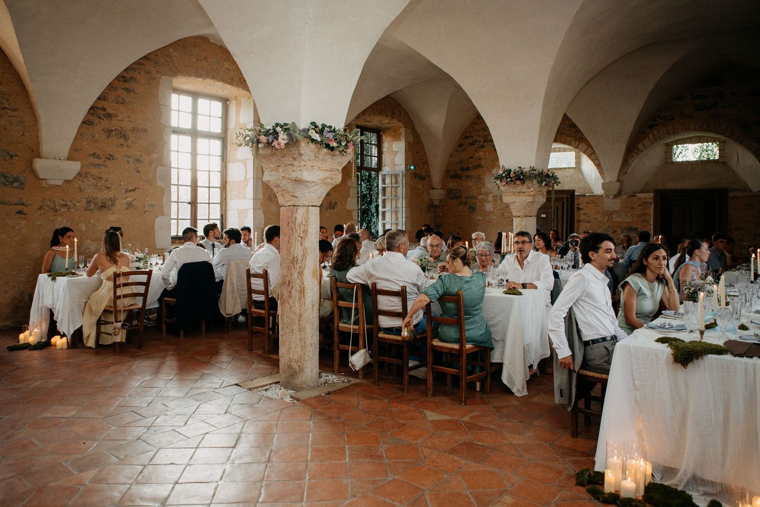 Elegant wedding reception in a rustic stone venue with arched ceilings, candlelit tables, and guests dressed in white attire.