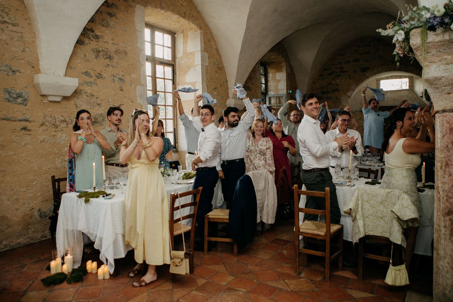 Wedding guests celebrating in a rustic stone venue with arched ceilings, raising glasses and cheering at a reception dinner.