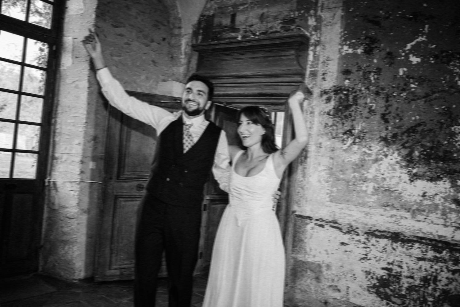 Black and white photo of a joyful couple in wedding attire celebrating indoors near rustic stone walls.
