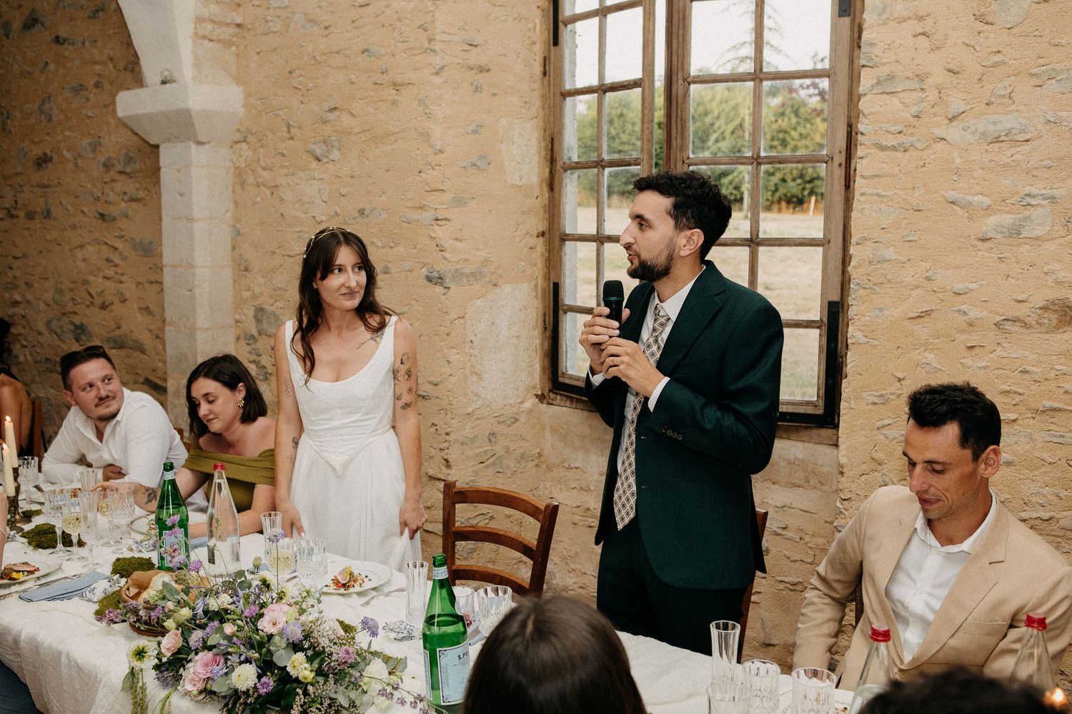 A groom gives a speech at an elegant wedding reception dinner in a rustic stone venue with floral centerpieces.