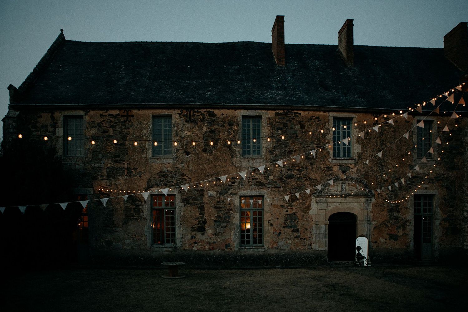 Historic stone manor adorned with fairy lights and bunting at dusk, creating a magical wedding venue atmosphere.
