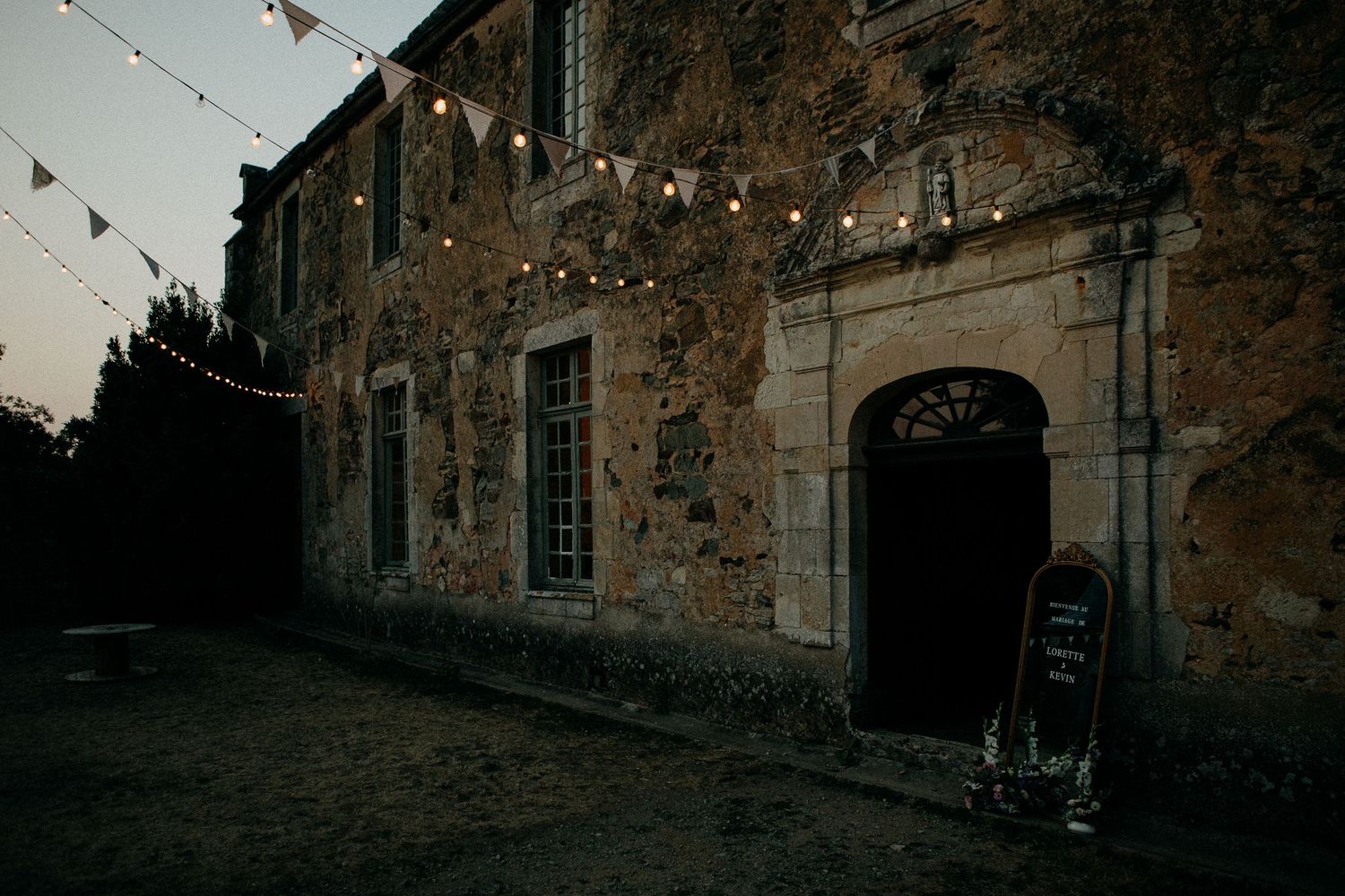 Rustic stone building at dusk with string lights and bunting decorations, featuring an arched doorway and chalkboard sign.