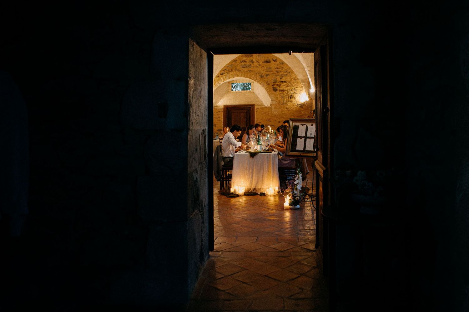 Elegant wedding reception viewed through dark doorway, guests gathered around candlelit table in vaulted stone venue.