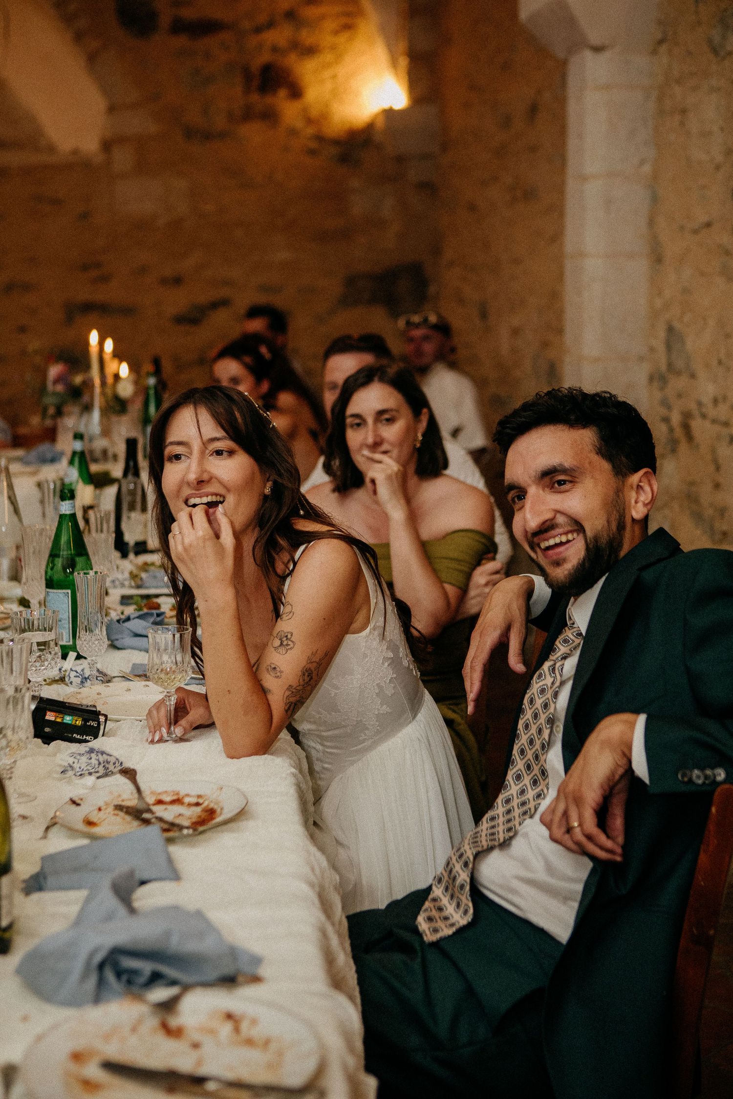 Bride and groom laughing joyfully at wedding reception dinner table in rustic stone venue with candles.