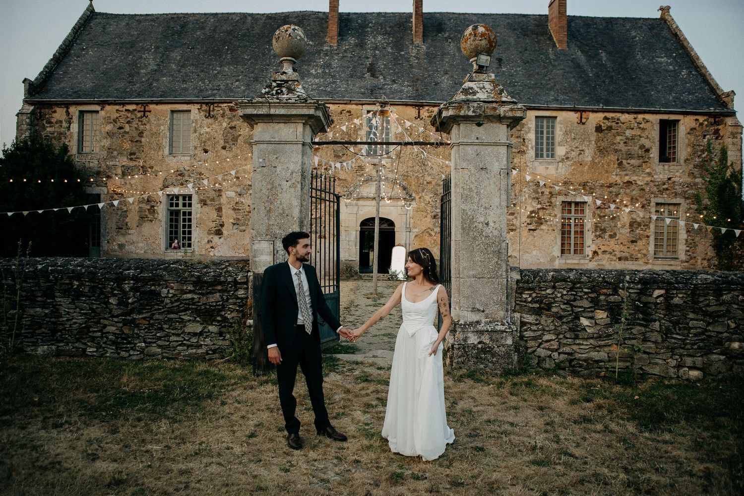 Bride and groom hold hands outside a rustic stone French chateau adorned with fairy lights at dusk.