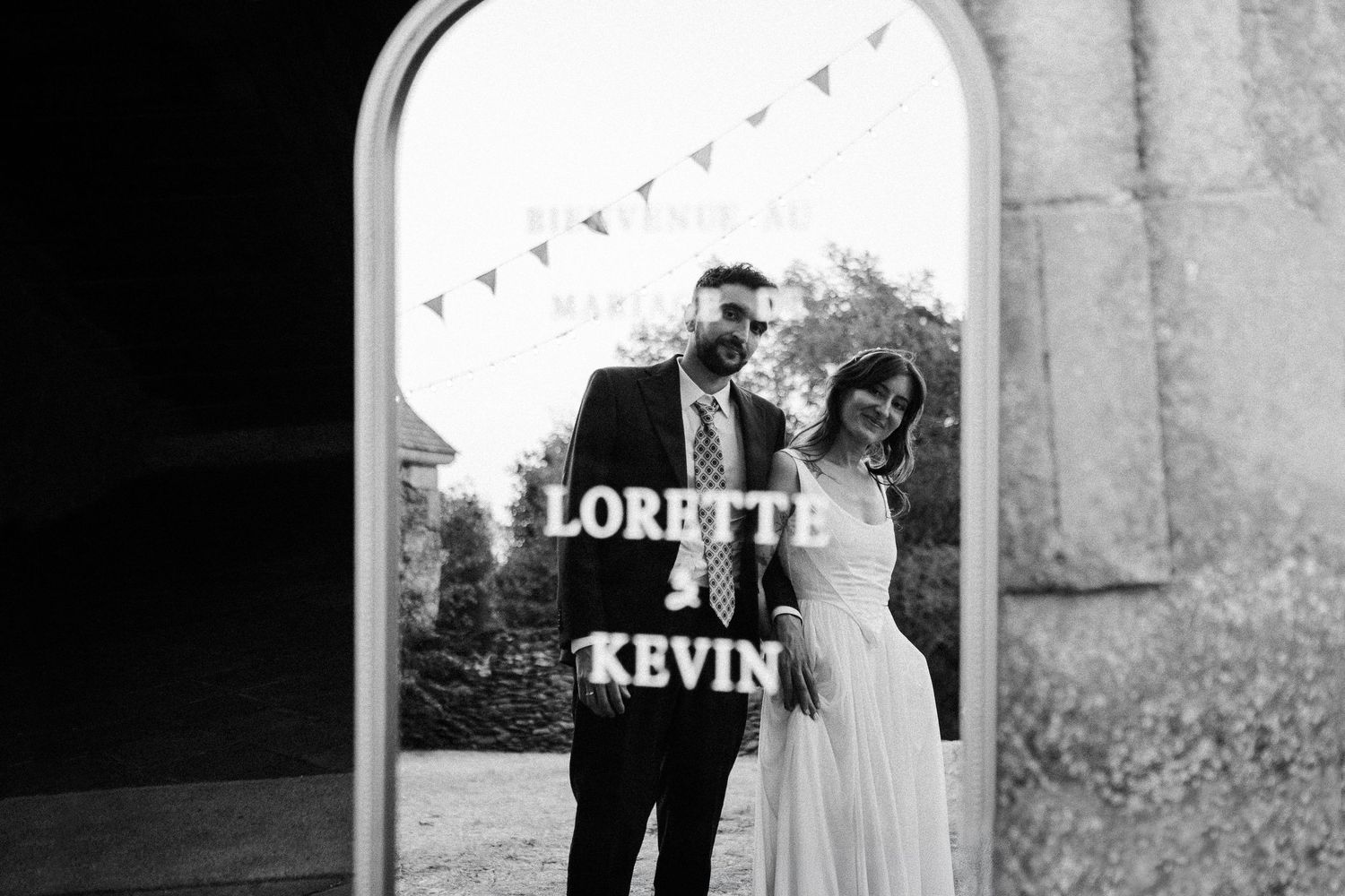 Black and white photo of bride and groom posing behind a mirror sign reading Lorette and Kevin at their wedding.