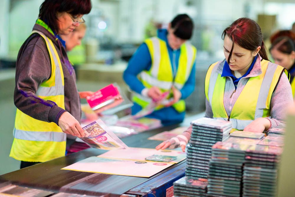 Workers in yellow safety vests operating machinery, sorting and packaging printed materials on a production line in a warehouse.