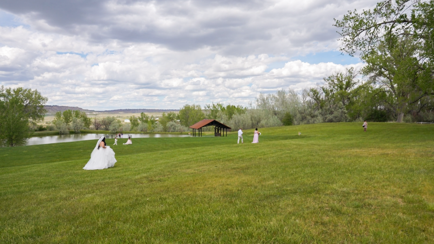 Outdoor wedding ceremony on lush green lawn with bridal party in white dresses near a pond and red barn.