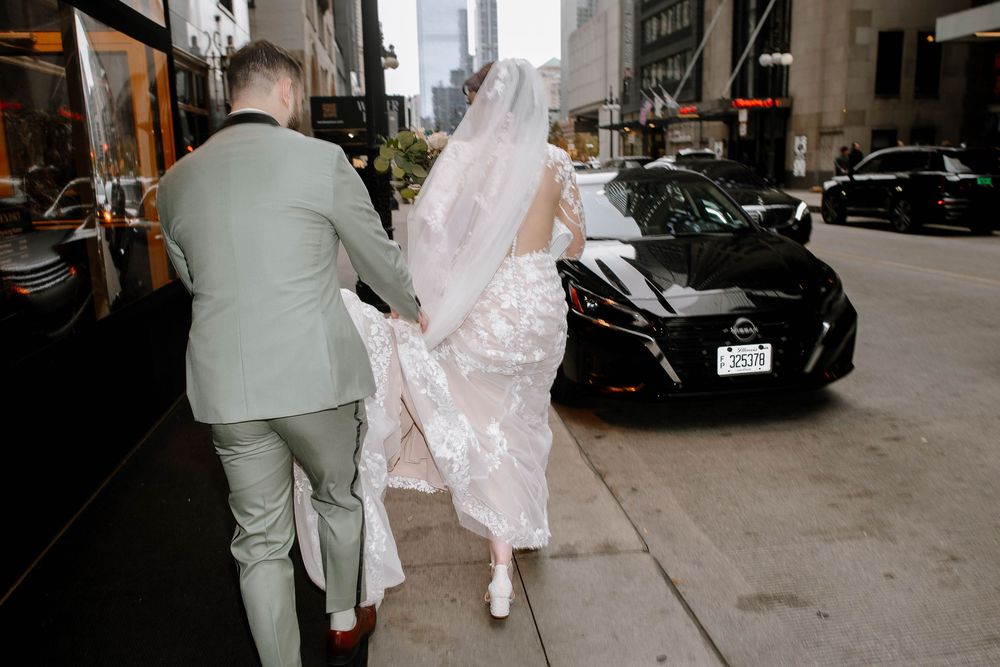 Bride and groom walking hand-in-hand on a city sidewalk, bride in white lace gown with flowing veil.