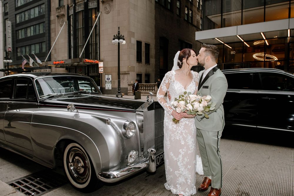 Bride and groom kissing beside a vintage silver Rolls-Royce on a city street, bride holding white bouquet.