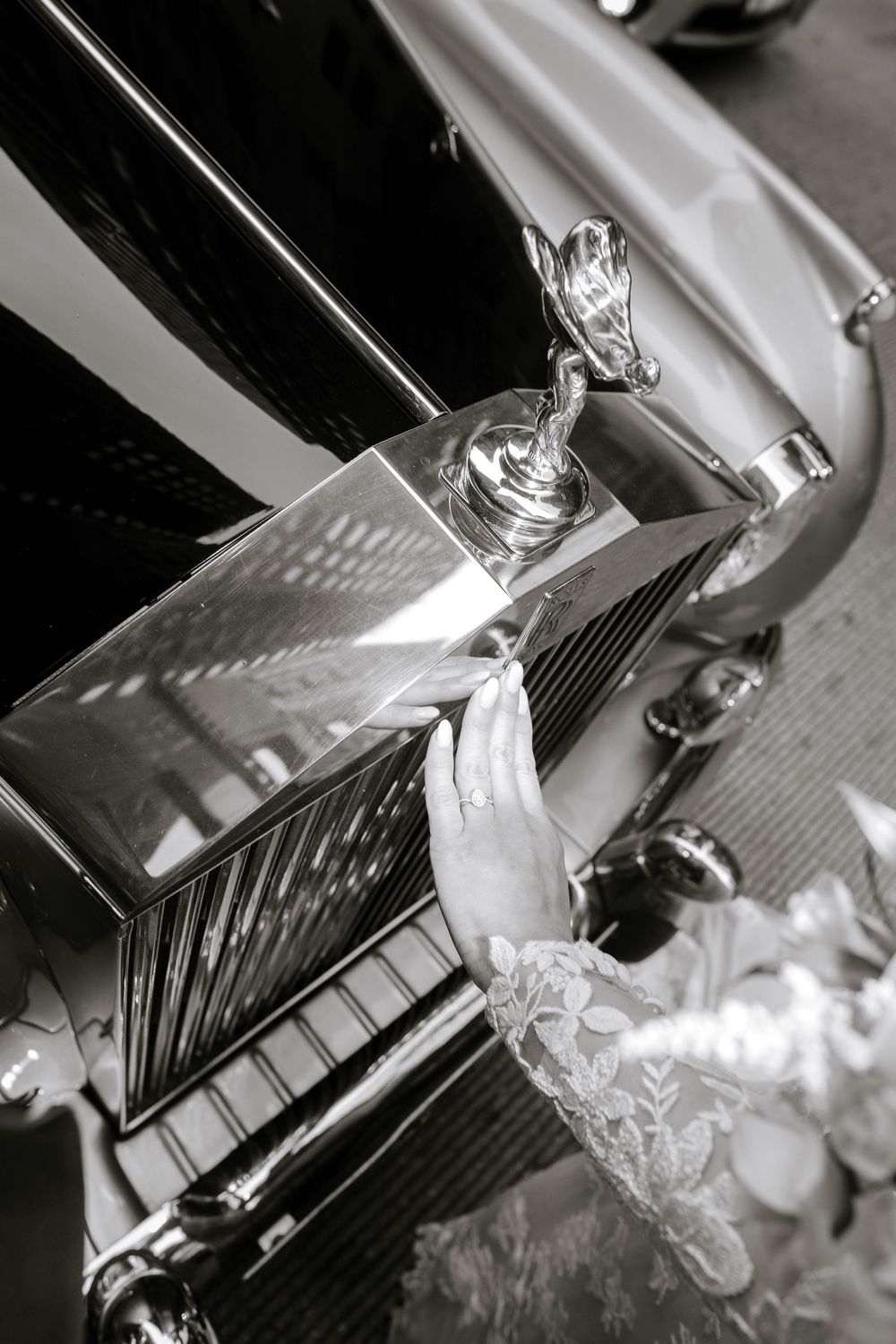 Black and white photo of a bride's lace-gloved hand touching a vintage Rolls-Royce hood ornament and chrome grille.