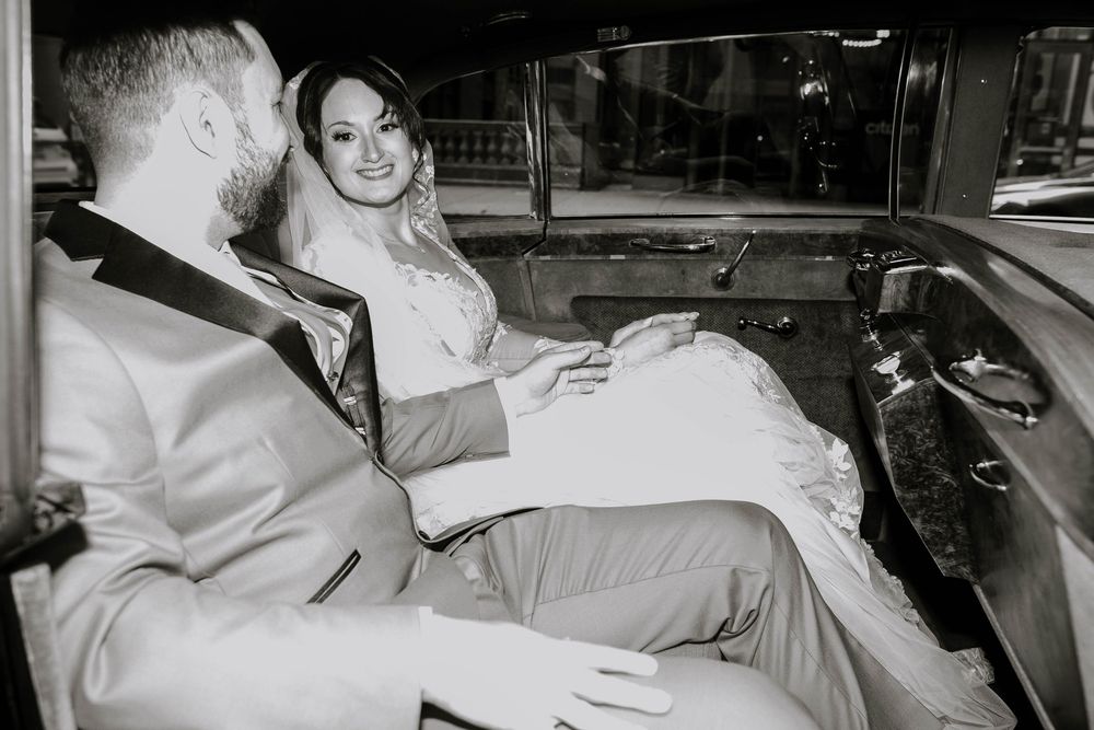Black and white photo of a bride and groom sharing a romantic moment inside a classic vintage car on their wedding day.
