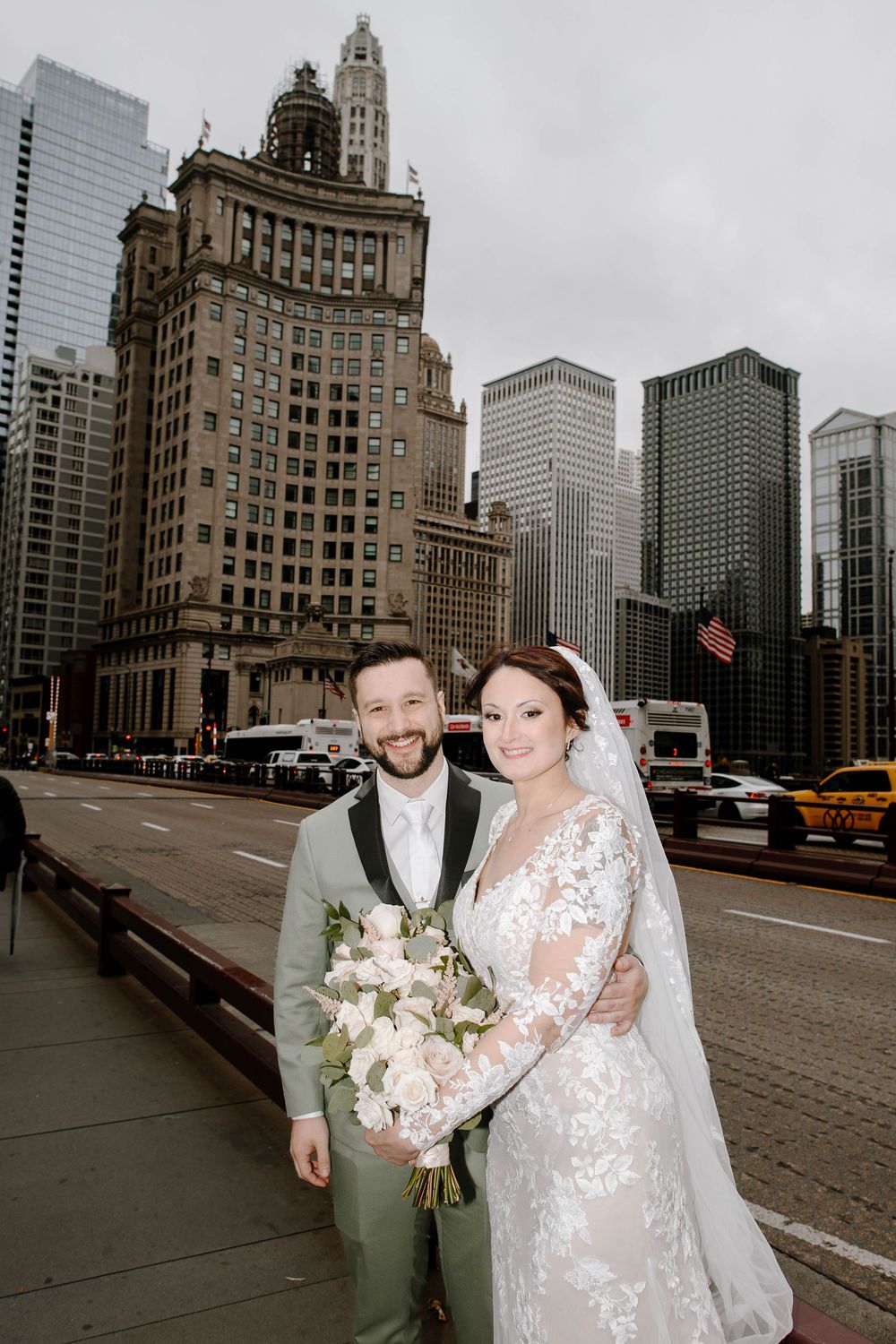 Bride and groom pose on a Chicago bridge with city skyline and historic buildings in the background on their wedding day.