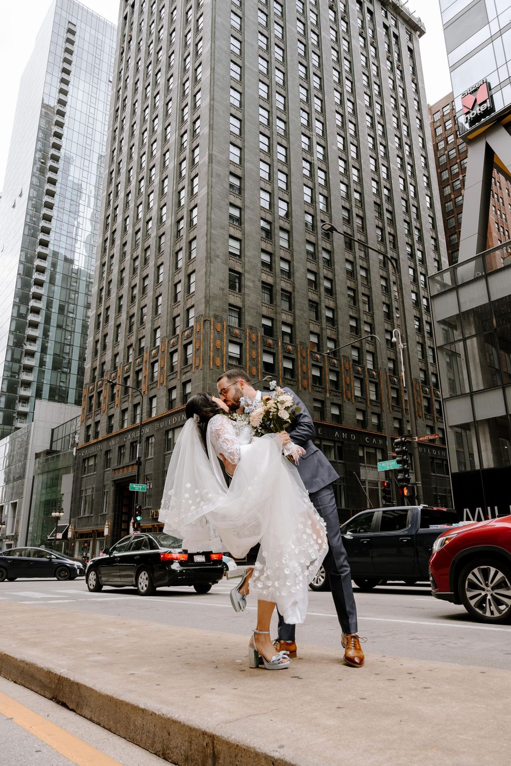 Bride and groom share a romantic dip kiss on a city street surrounded by tall buildings and urban traffic.