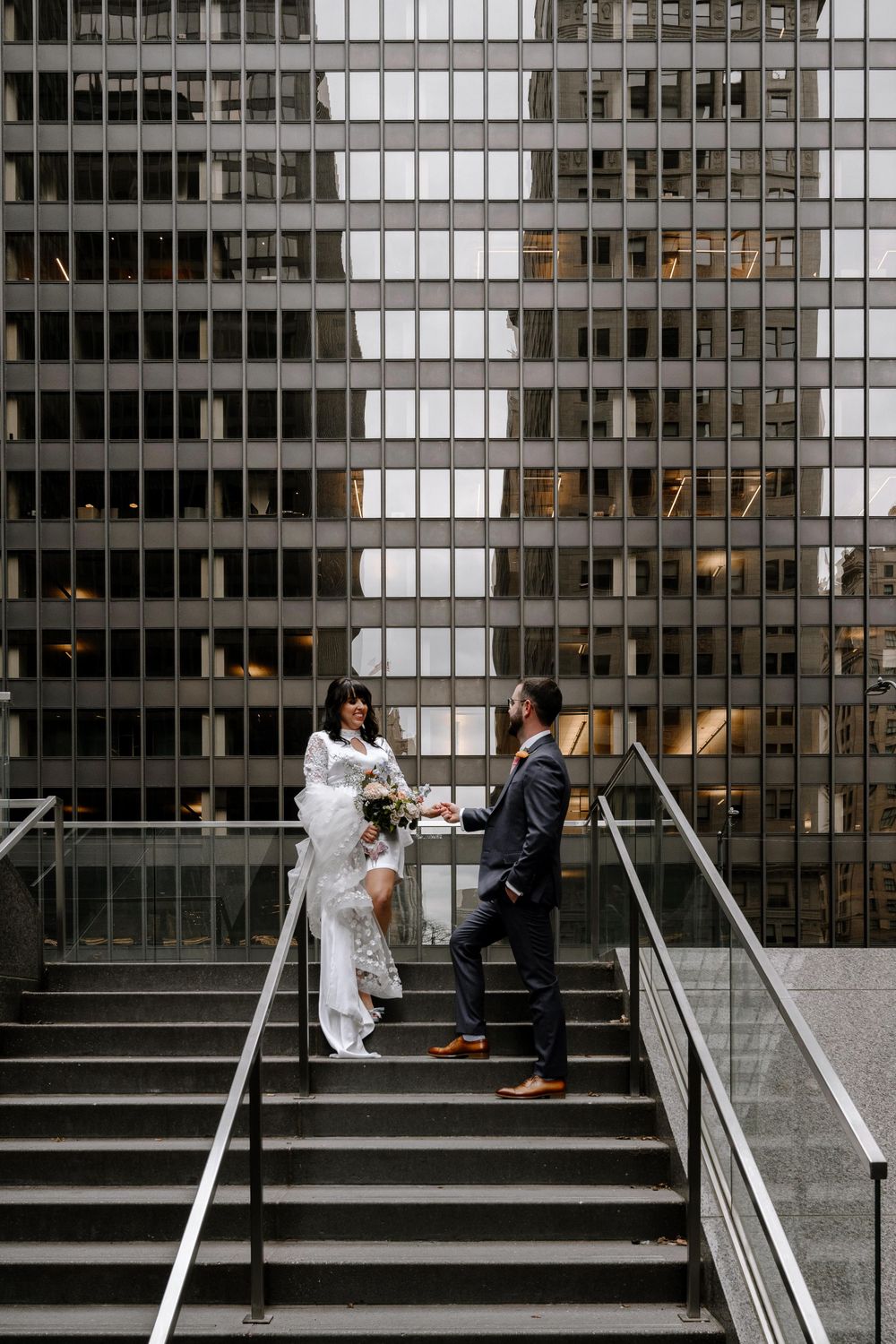 Bride and groom holding hands on urban stairs with modern glass skyscraper backdrop during city wedding photoshoot.
