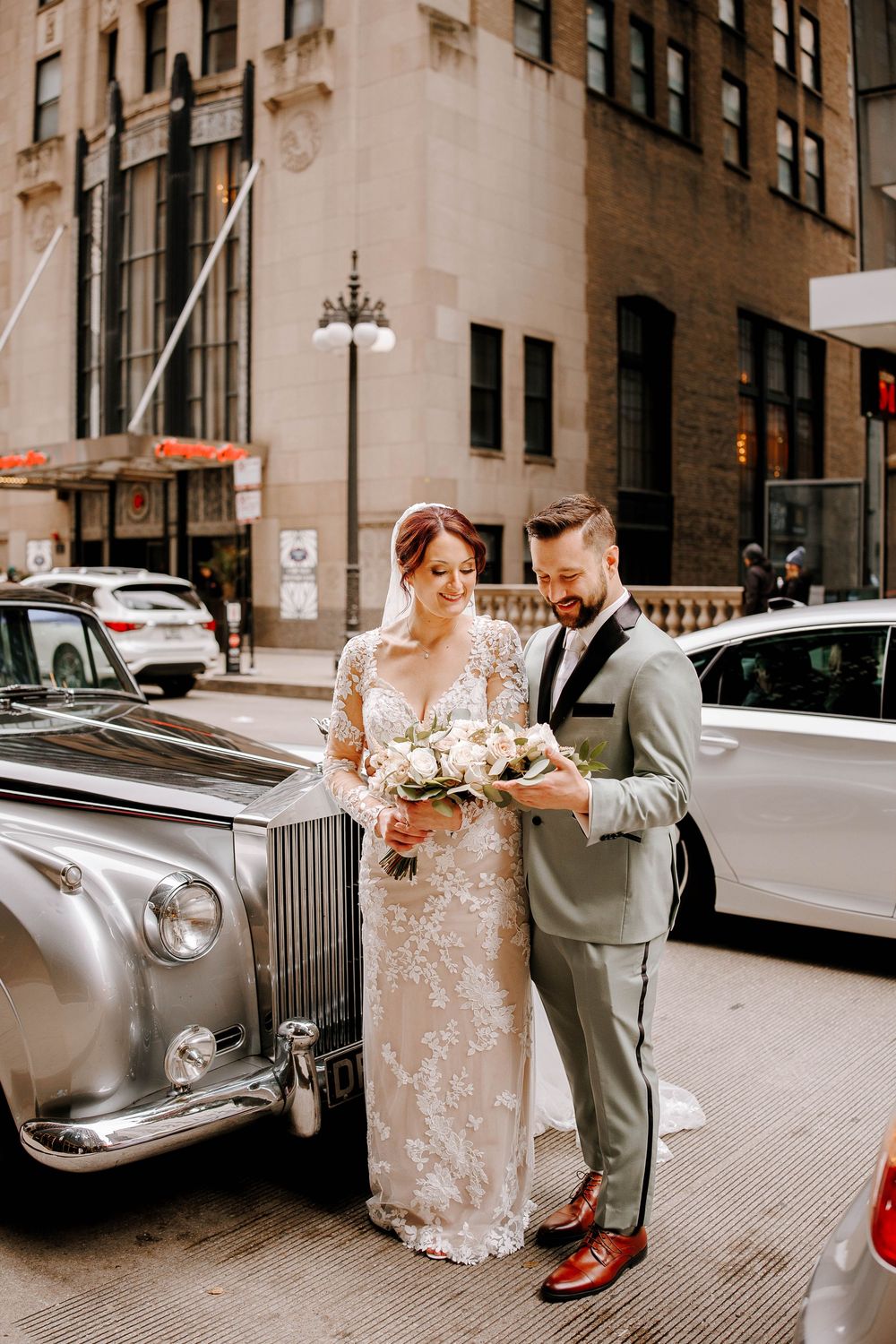 Bride and groom pose beside a vintage silver Rolls-Royce on a city street, bride holding a white floral bouquet.