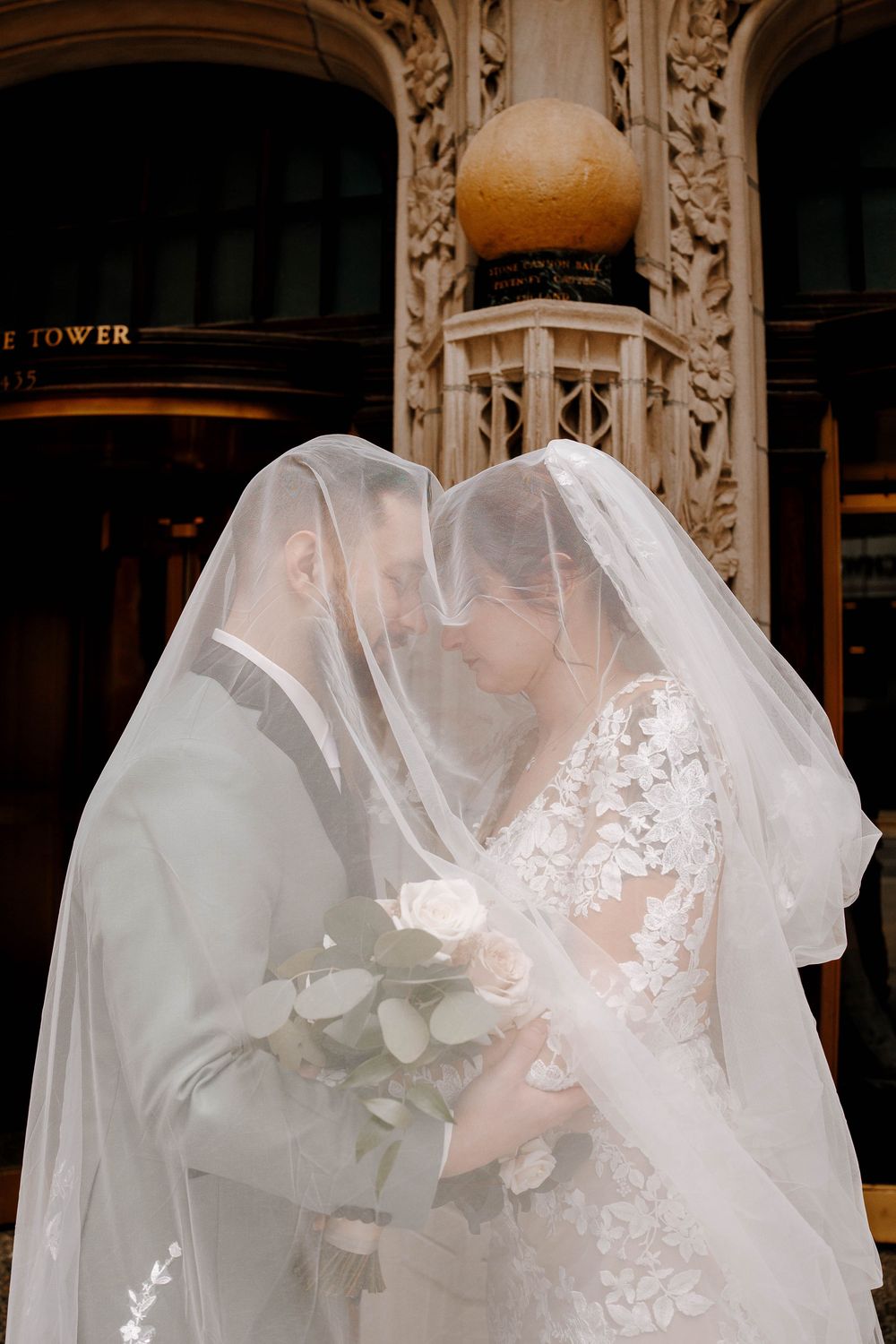 Bride and groom share an intimate moment under a flowing white veil, bride holding white roses, outside an ornate building.