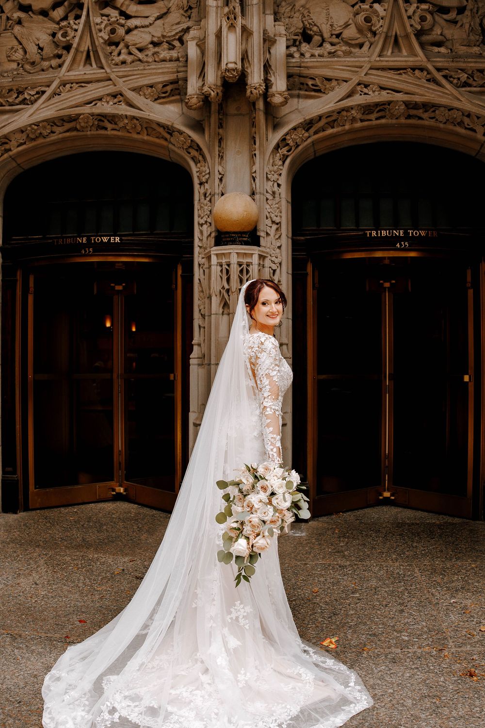 Bride in lace gown with long veil holds white bouquet outside Tribune Tower in Chicago.