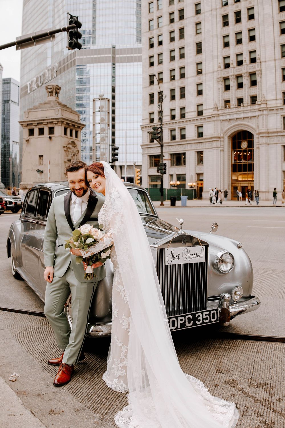 Bride and groom pose by a vintage Rolls-Royce Just Married car on a city street with tall buildings in Chicago.
