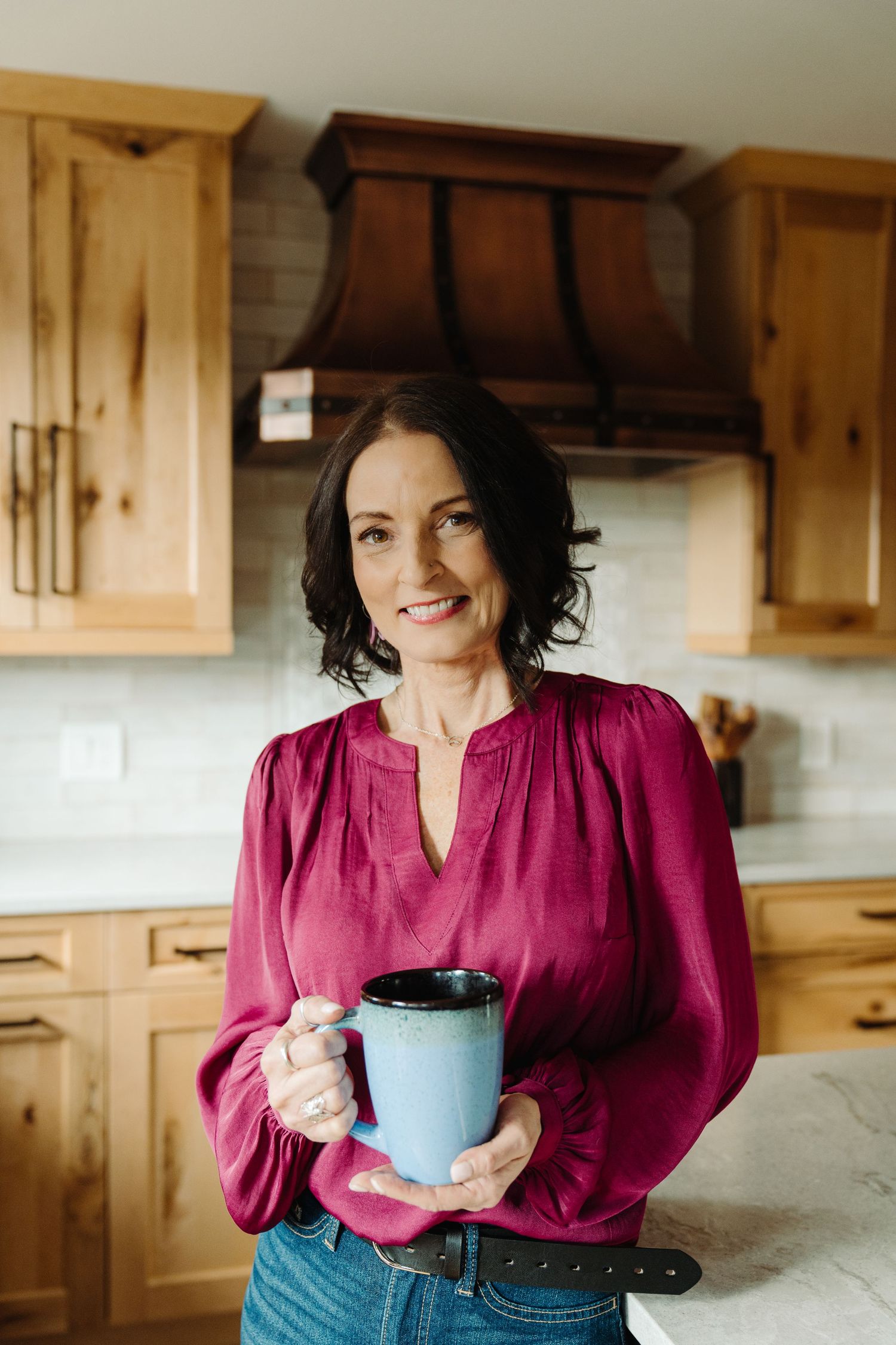 Woman in magenta blouse holding blue mug smiling in modern kitchen with wood cabinets.