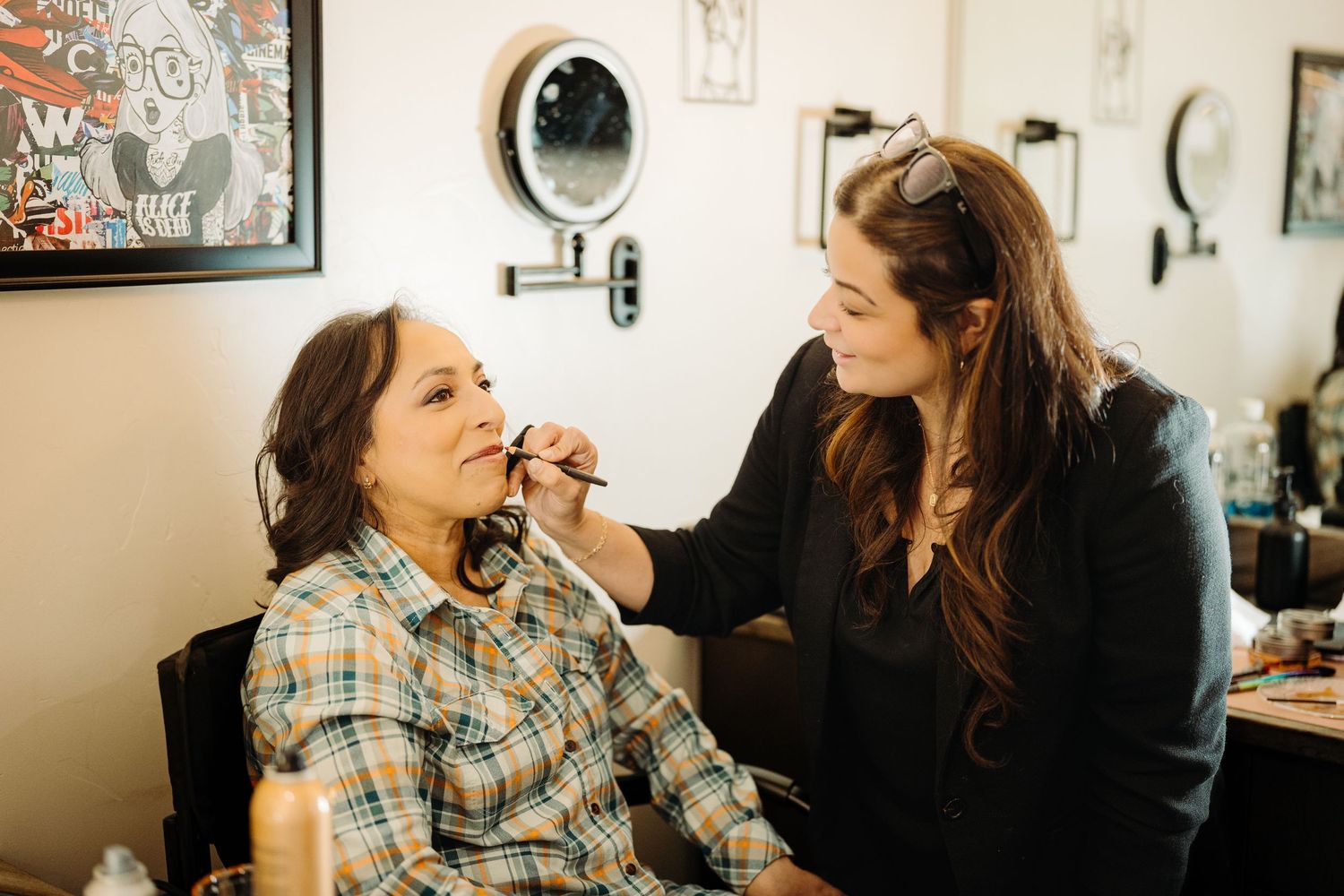 A makeup artist applies makeup to a seated woman in a beauty salon with mirrors and artwork on the walls.