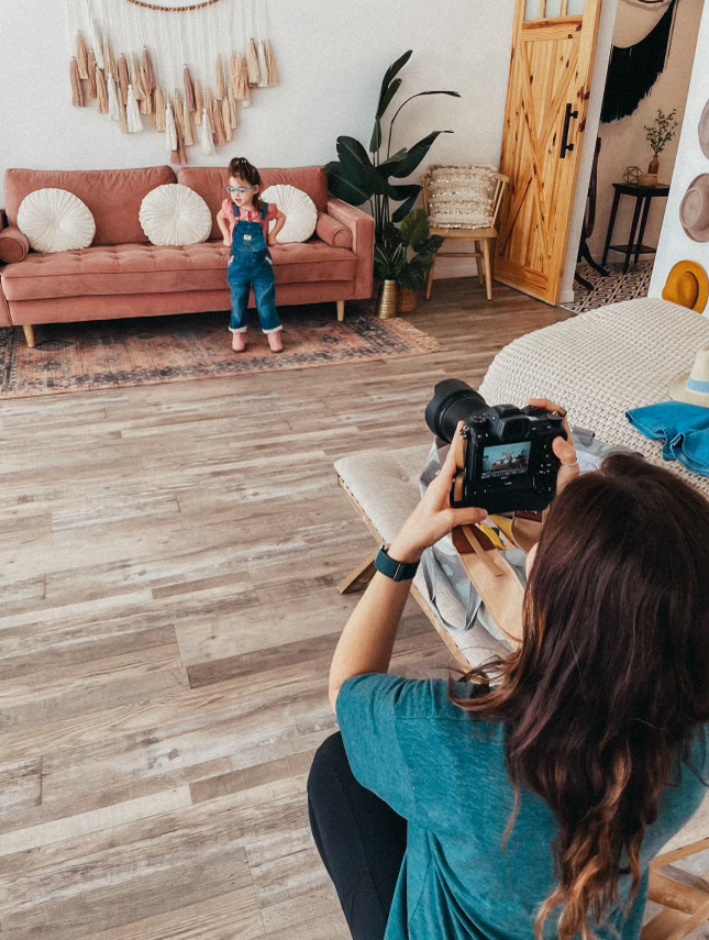 Photographer capturing a girl in blue overalls standing in a boho-styled living room with pink sofa.