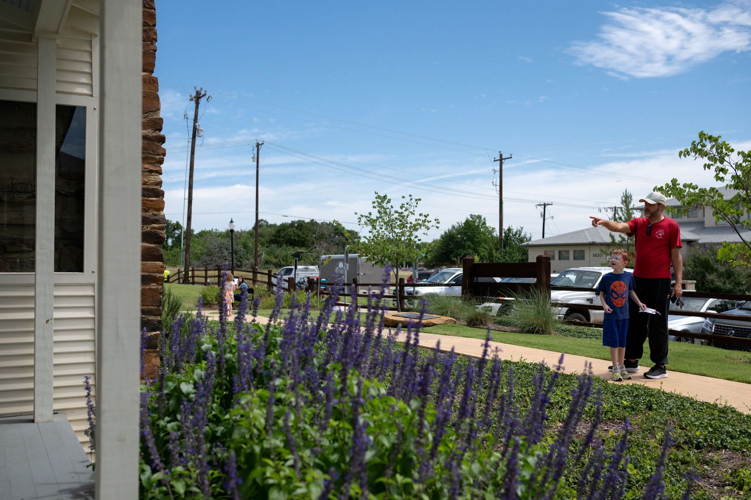 Adult and child explore a garden path with purple salvia flowers near a historic building on a sunny day.