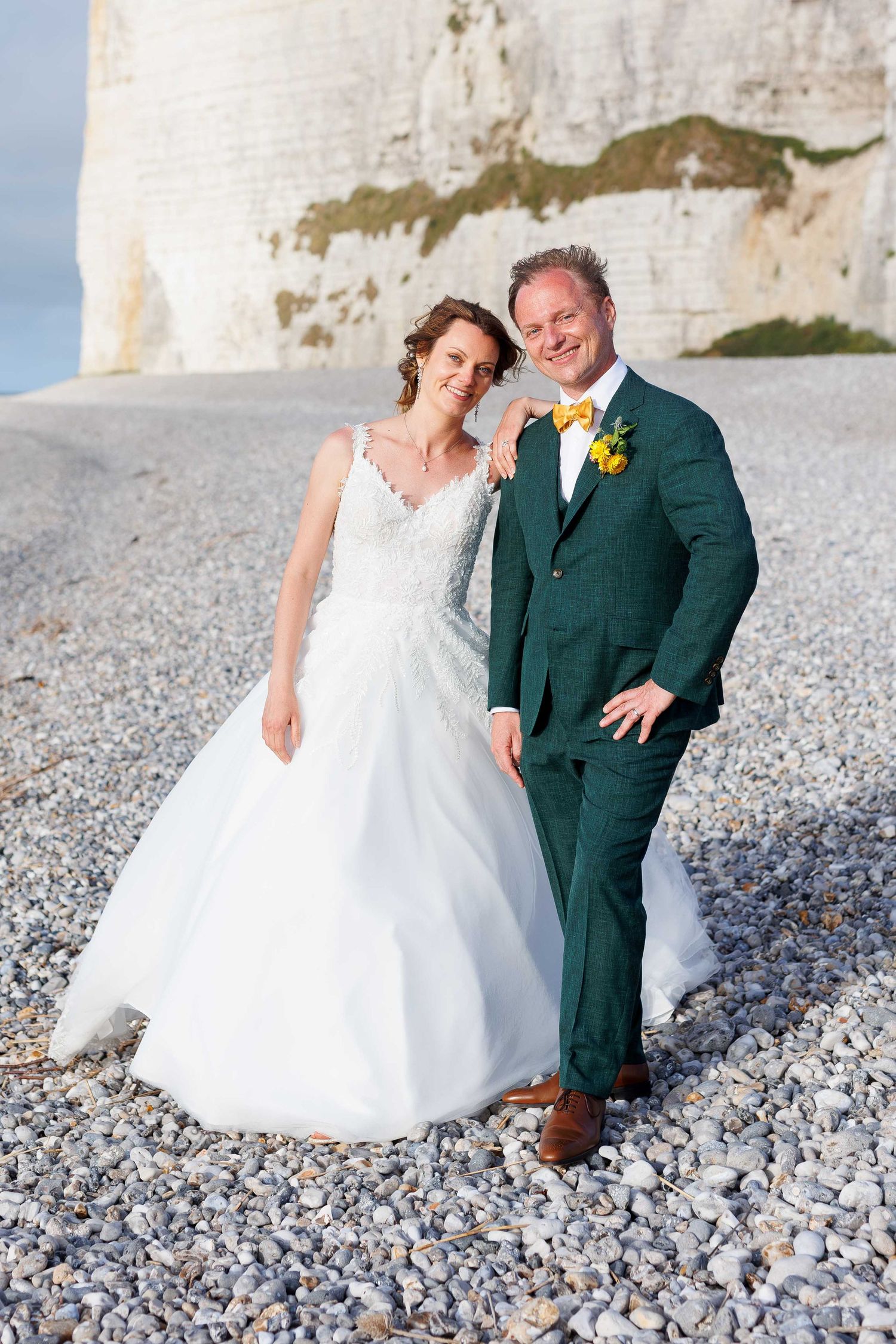 Bride in white gown and groom in green suit with yellow bow tie pose on pebble beach with white cliffs behind.